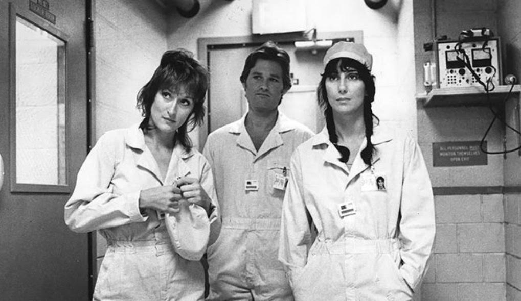 A black & white image of Meryl Streep, Kurt Russell & Cher wearing jumpsuits inside a lab.