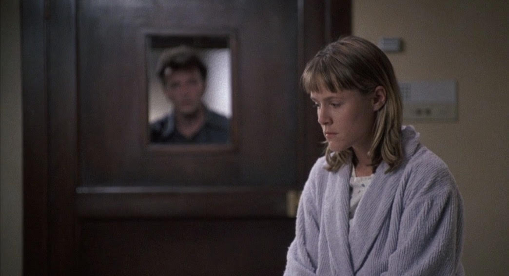 Joon is a white woman in a locked hospital room. Her brother Benny watches her through the window on the door, looking concerned.