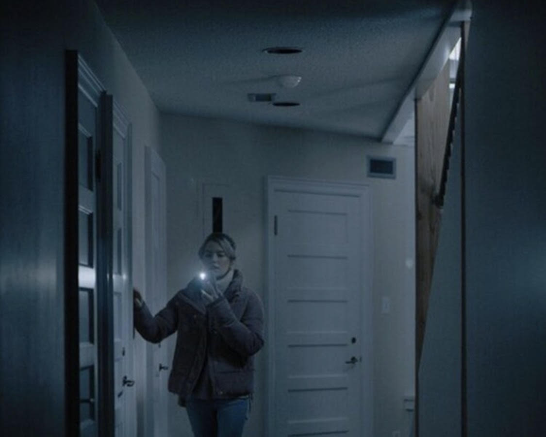 A white woman in a dark hallway holds her phone up in front of her with the flashlight shining forward as her hand holds the wall.