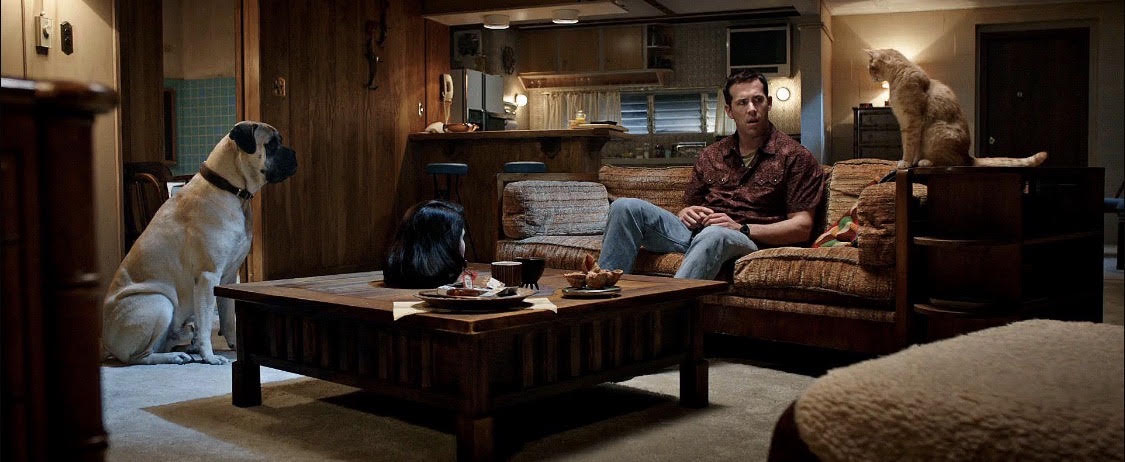 Ryan Reynolds sits on a couch with his cat sitting on the couch near him and his dog on the floor looking right at him.