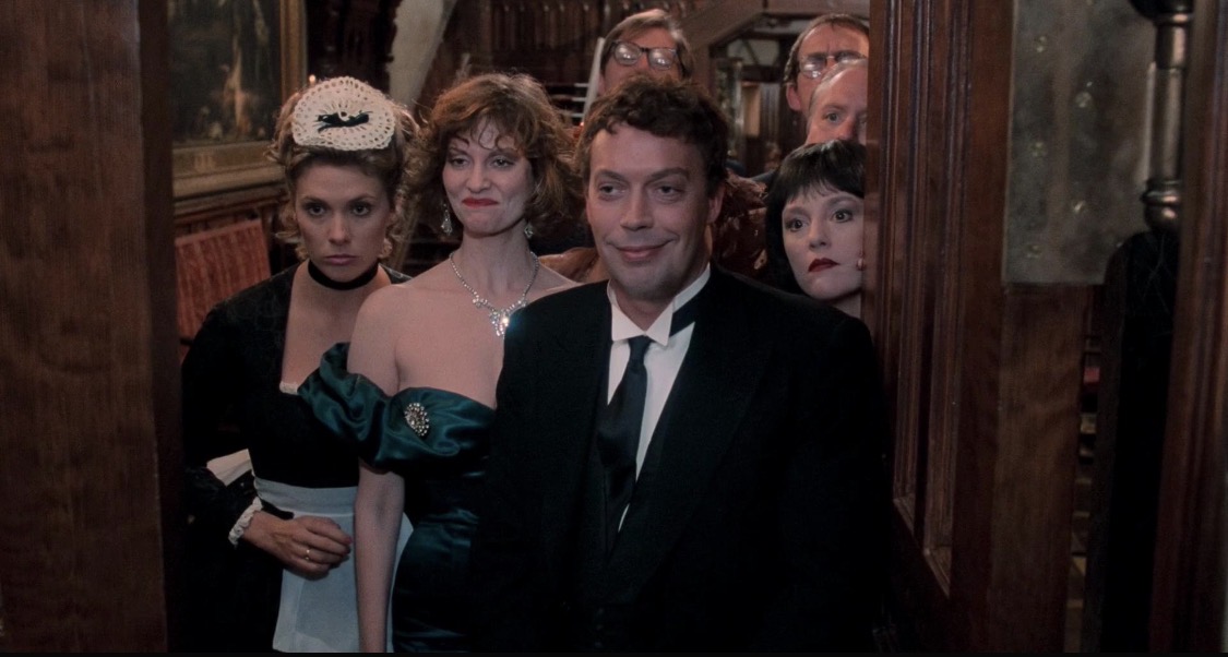 Clue: Tim Curry stands in doorway with other cast members behind him looking at the newly arrived guest